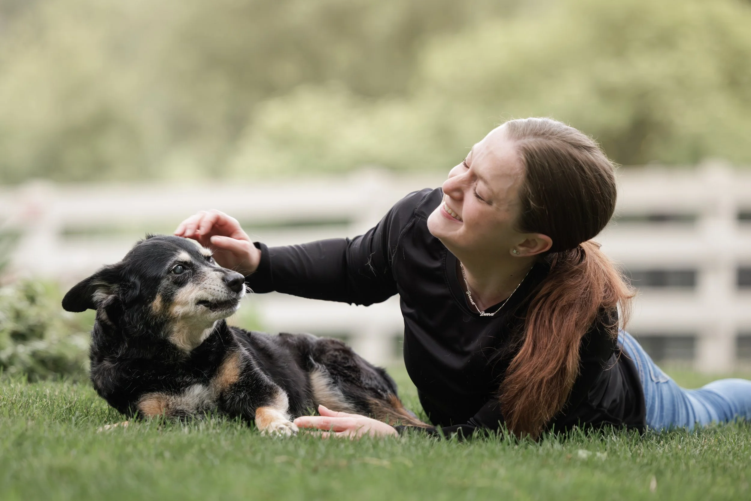 A woman lying on the grass petting a dog, smiling in an outdoor setting with a white fence and trees in the background.