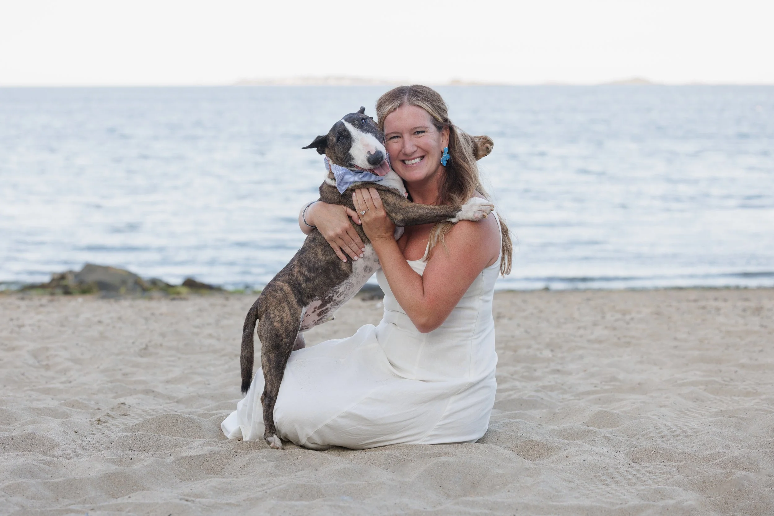 Woman in white dress smiling while sitting on sandy beach, hugging a brindle and white dog with a bow tie, near the water with distant land in the background.