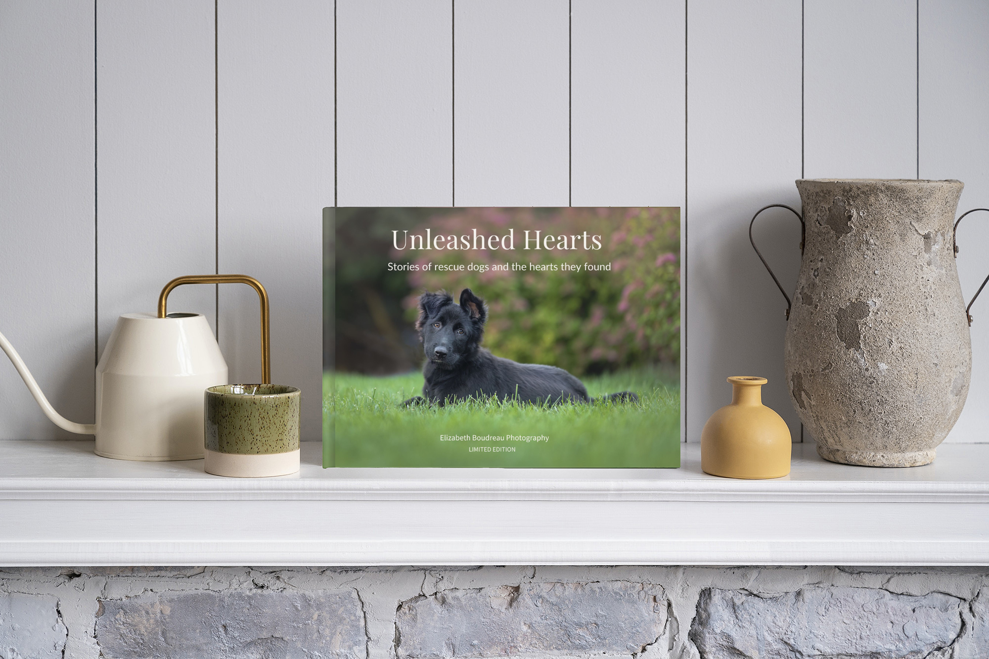 A book titled 'Unleashed Hearts' featuring a black dog lying on grass with pink flowers in the background, placed on a white mantel with various vases and a white wall with vertical paneling behind it.